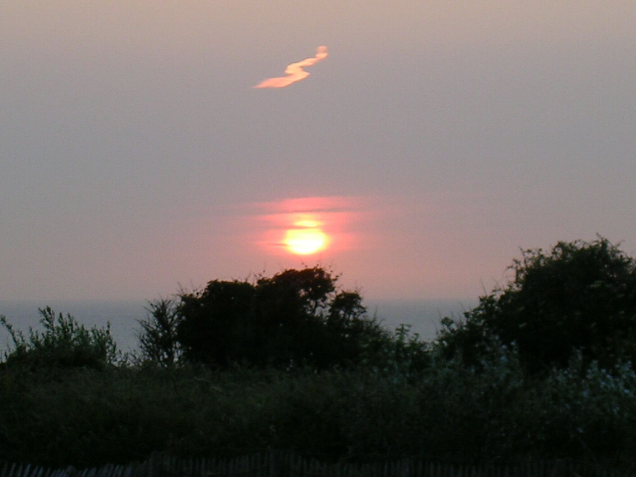 A sunset with the sun partially obscured by clouds, silhouetted trees, and a fence in the foreground.