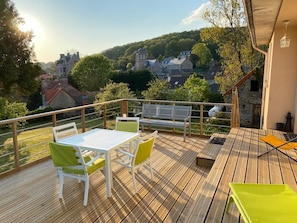 Une terrasse en bois avec une table et des chaises, d’où l’on a vue sur un village avec ses bâtiments et ses arbres.