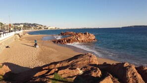Una playa con un promontorio rocoso, una persona pescando y un paisaje urbano distante.