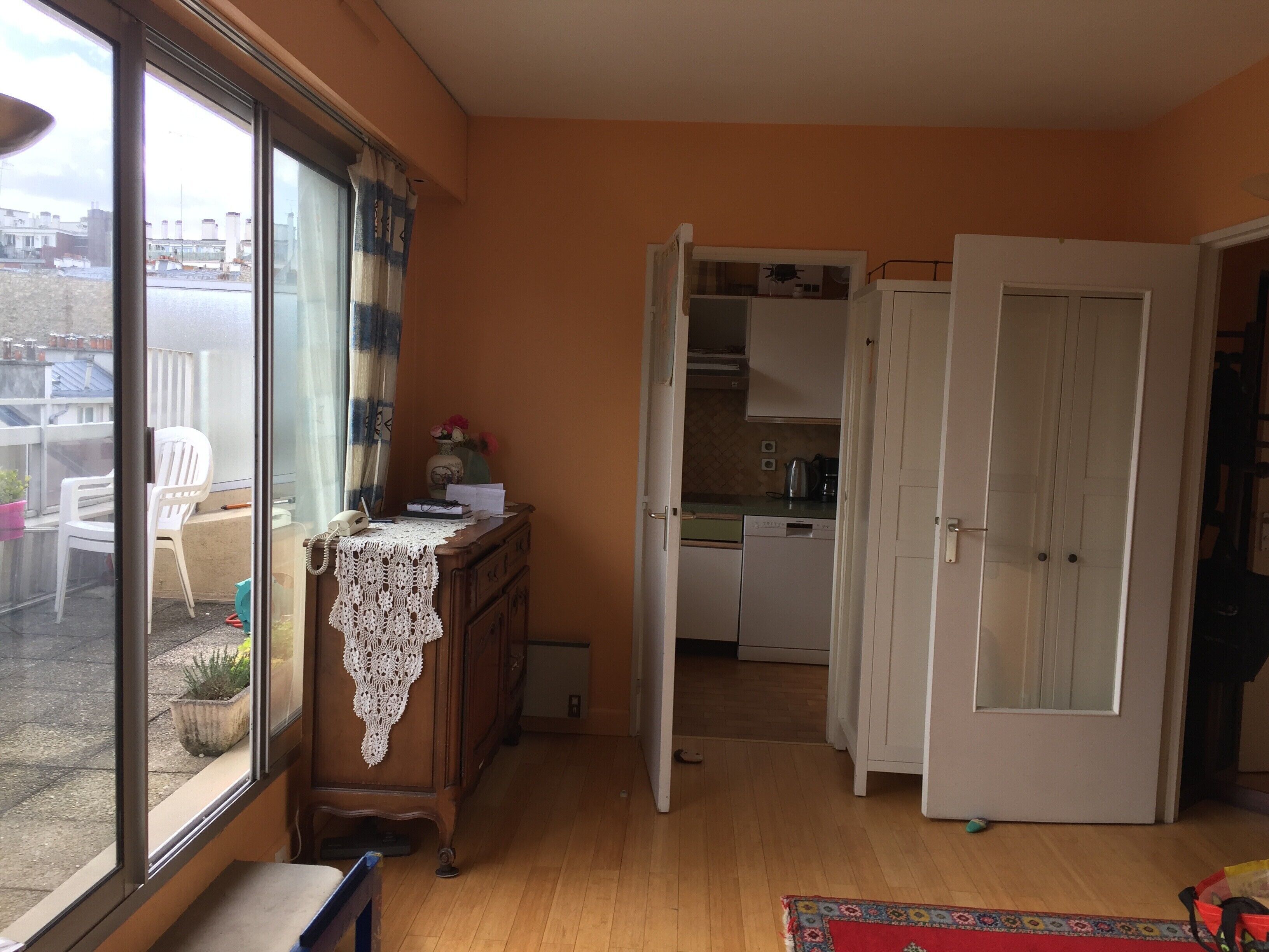 Une pièce comprenant une commode en bois, une petite table recouverte d’une nappe en dentelle, une chaise et une vue sur un balcon agrémenté d’une chaise et d’un pot de fleurs.