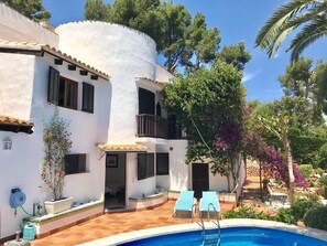 Eine weiße Villa mit Pool, zwei Liegestühlen und einem Garten mit Bäumen und Blumen.