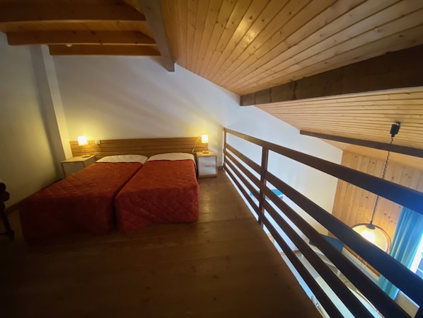 Une chambre avec deux lits, des poutres apparentes en bois au plafond et une rambarde de balcon.