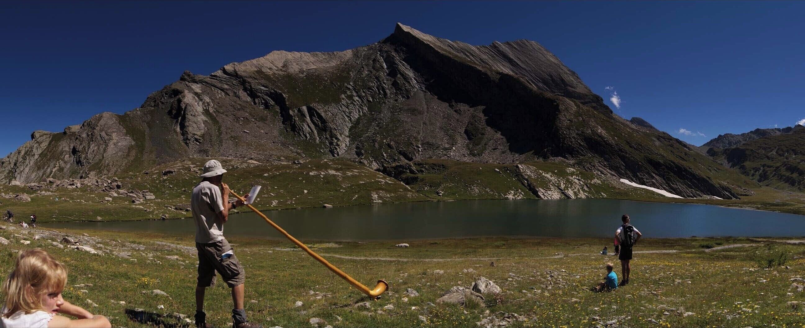 Eine Berglandschaft mit einem See, einer Person mit Wanderstock und einer anderen Person, die auf dem Gras sitzt.
