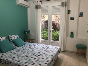 Et soveværelse med en seng, turkise puder, et natbord, en lampe, et vindue med gardiner og en lille skammel.