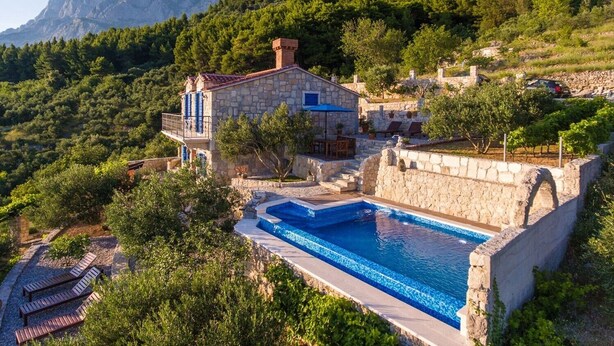 A stone villa with a swimming pool, surrounded by lush greenery and mountains.