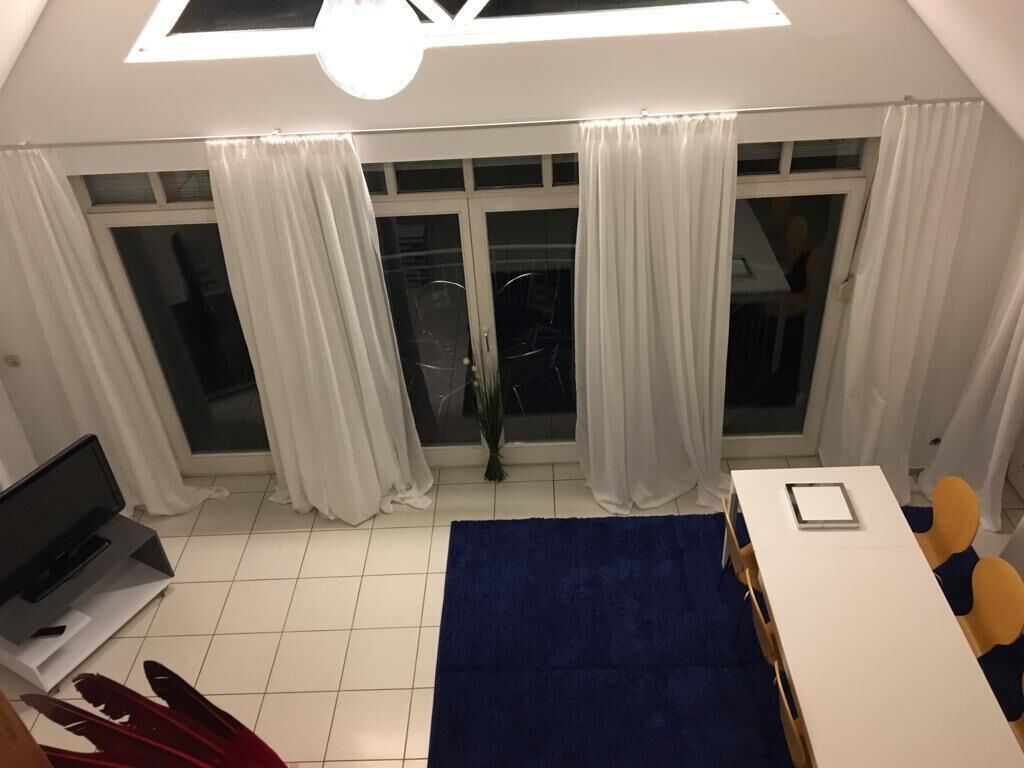 Quarto com uma grande janela, cortinas brancas, um tapete azul, uma mesa branca e uma cadeira.