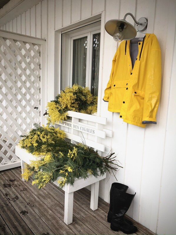 Um banco de madeira branca com flores amarelas e vegetação, um casaco amarelo pendurado na parede e uma bota preta no chão.