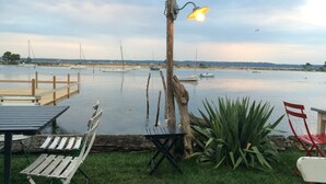 A waterfront patio with wooden tables and chairs, a lamp post, and a view of boats on the water.