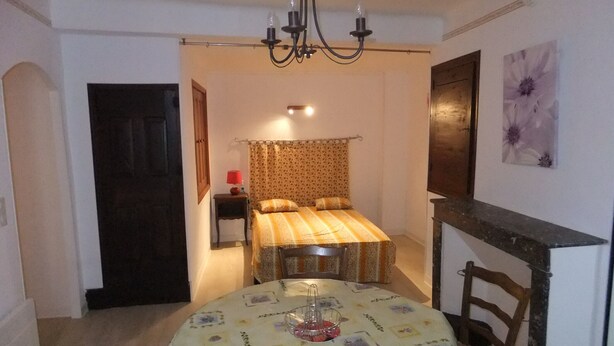 Une pièce chaleureuse avec un lit, une table recouverte d’une nappe à motifs floraux, un lustre et un tableau accroché au mur.