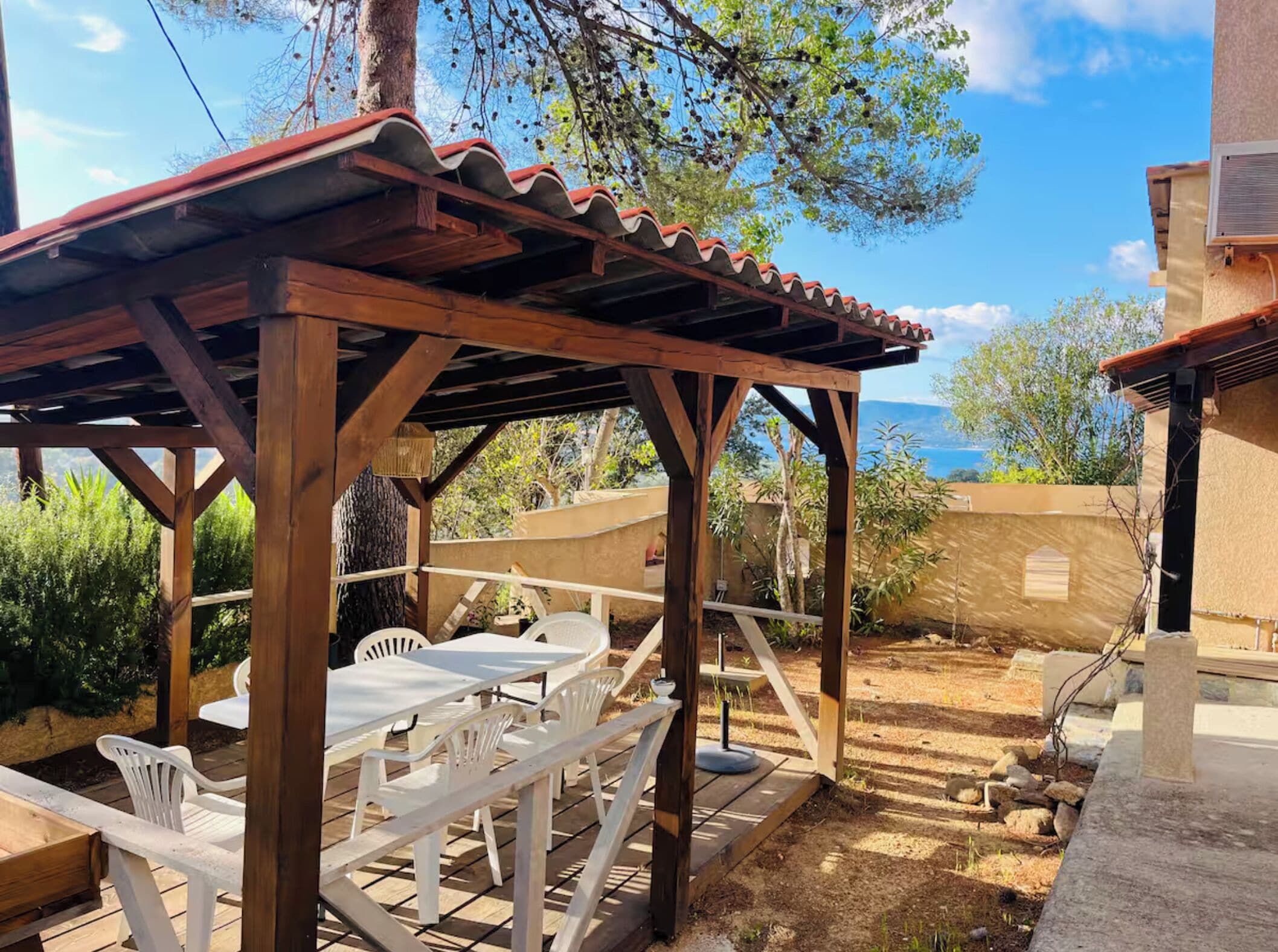 A wooden pergola with a tiled roof, white plastic chairs, and a table set up for outdoor dining.