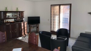Ein Wohnzimmer mit einem Holzesstisch, zwei schwarzen Sesseln, einem Sofa, einem Fernseher auf einem Schrank und einem großen Fenster mit Vorhängen.