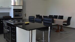 Een moderne keuken met een gasfornuis, oven en roestvrijstalen kasten. Er is een eethoek met een tafel en stoelen.
