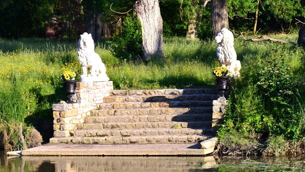 Un escalier en pierre mène à un petit promontoire orné de statues de lions, le tout étant entouré d’une végétation luxuriante et surplombant un plan d’eau paisible dans lequel se reflètent les statues.