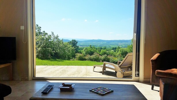 A room with a large window offering a view of a green landscape and mountains. There is a wooden table with a remote control and a book. - Very nice Mazet with pool and panoramic views of the Cevennes (Saint-Privat-de-Champclos)