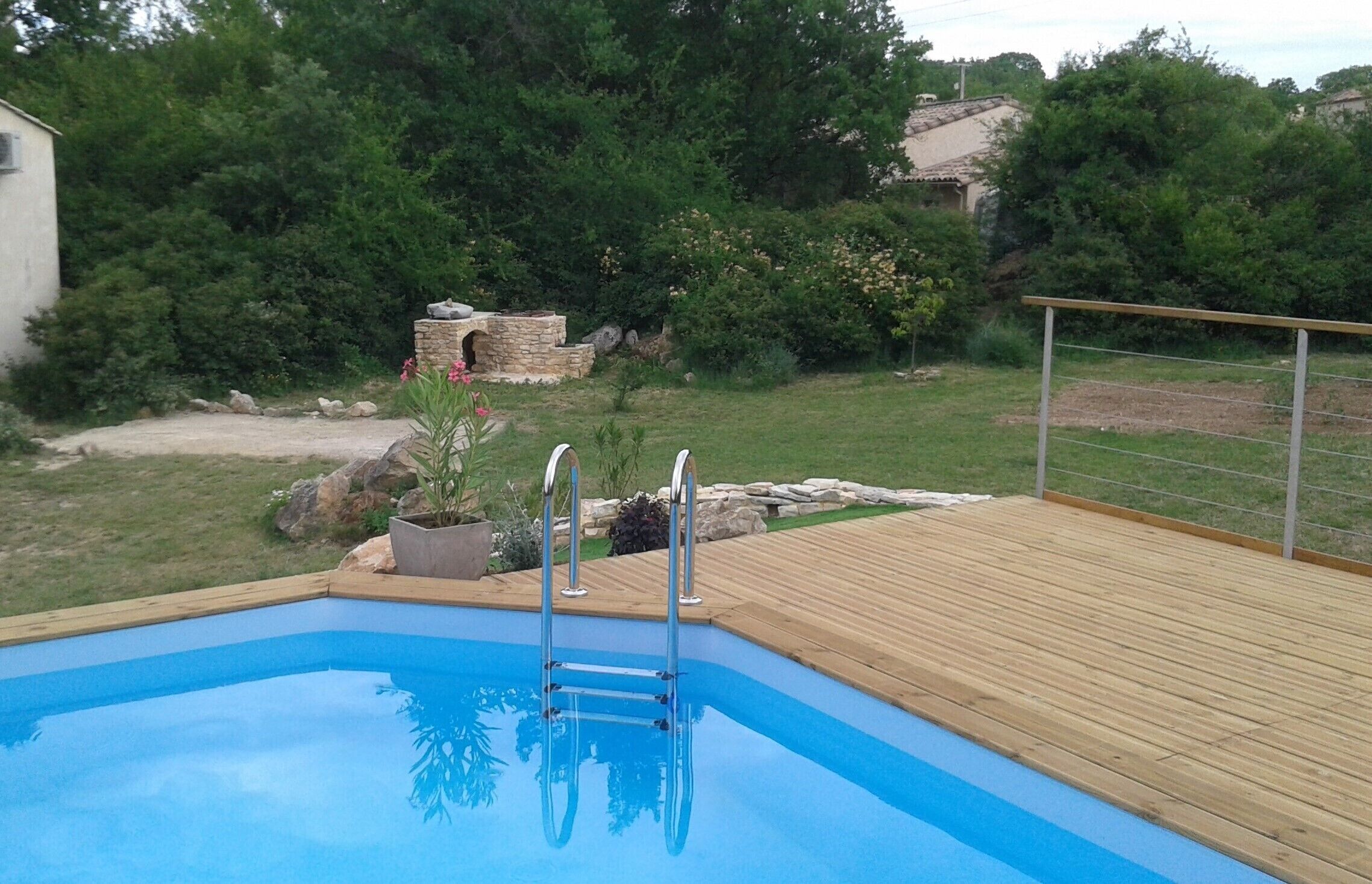 Ein Schwimmbad mit Holzterrasse, umgeben von Grünpflanzen und einer Steinstruktur.