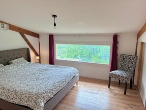 Un dormitorio con cama, ventana con vista a árboles, una silla y una lámpara.