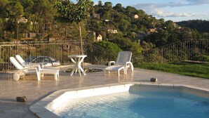 A swimming pool with lounge chairs and a table, surrounded by a metal fence, with a hillside and houses in the background.
