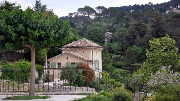 A two-story house with a tiled roof, surrounded by lush greenery and a metal fence.