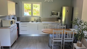 Een compacte keuken met een eethoek, witte kasten, een magnetron, een koelkast en een raam met uitzicht op groen.