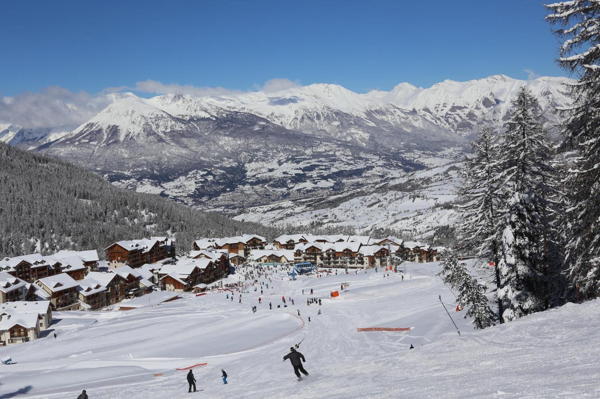 Un centro de esquí con varias cabañas de madera, una pista de esquí y montañas nevadas al fondo.