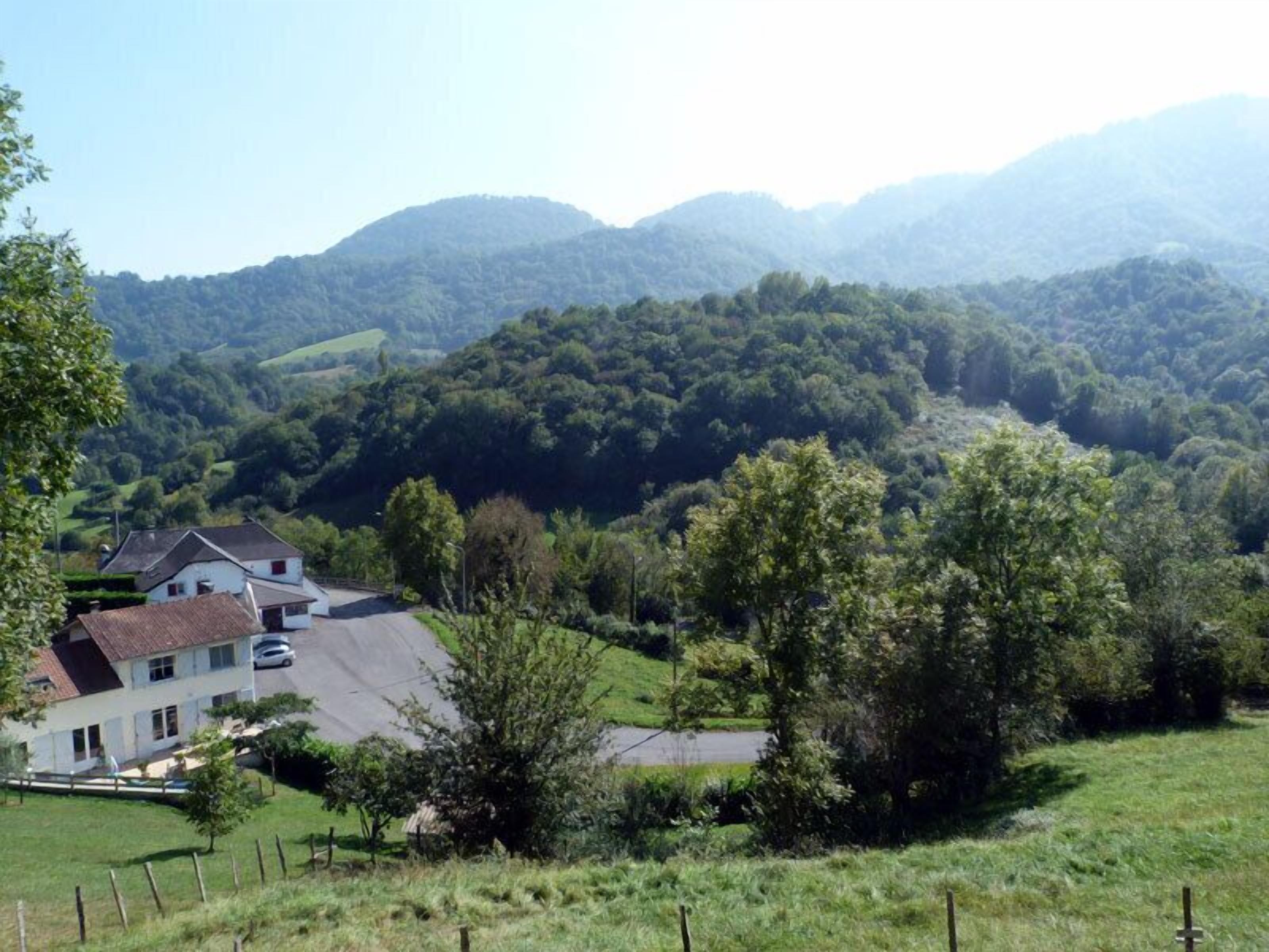 A rural house with a sloped roof, surrounded by lush greenery and hills in the background.