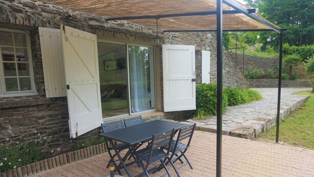 Een stenen gebouw met een overdekt terras, een tafel en stoelen, en een raam met luiken.