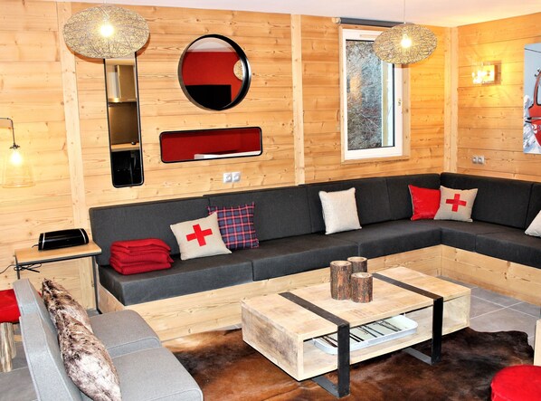 A modern living room with a wooden interior, a grey sofa with red and white cushions, a small table with a vase, and a large mirror on the wall.