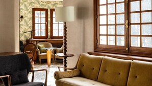 Ein gemütliches Wohnzimmer mit Holzbalkendecke, einem großen Fenster, einer Couch, einem Sessel und einem Couchtisch mit Muster.