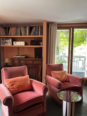 A room with two plush armchairs, a small table, a bookshelf with audio equipment, and a view of an outdoor area with chairs.