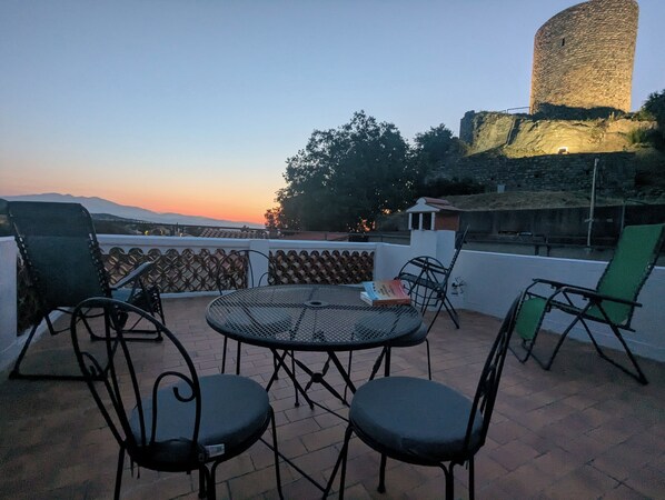 An outdoor seating area with a round table and chairs, overlooking a historic tower and distant mountains. - Charming house with garden and panoramic terrace; between sea and mountains! (Laroque-Des-Albères)