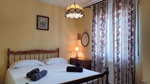 Un letto rifatto con lenzuola bianche, una testiera in legno e due asciugamani arrotolati. C'è un comodino con una lampada e un quadro incorniciato appeso al muro.