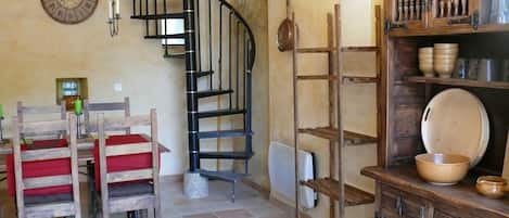 Una cocina con una escalera de caracol, sillas de madera, una mesa de comedor y estantes con utensilios de cocina.