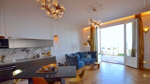 Une cuisine moderne avec un dosseret en marbre, un coin repas avec une table noire et des chaises orange, un canapé bleu et une grande porte-fenêtre coulissante donnant sur un balcon.