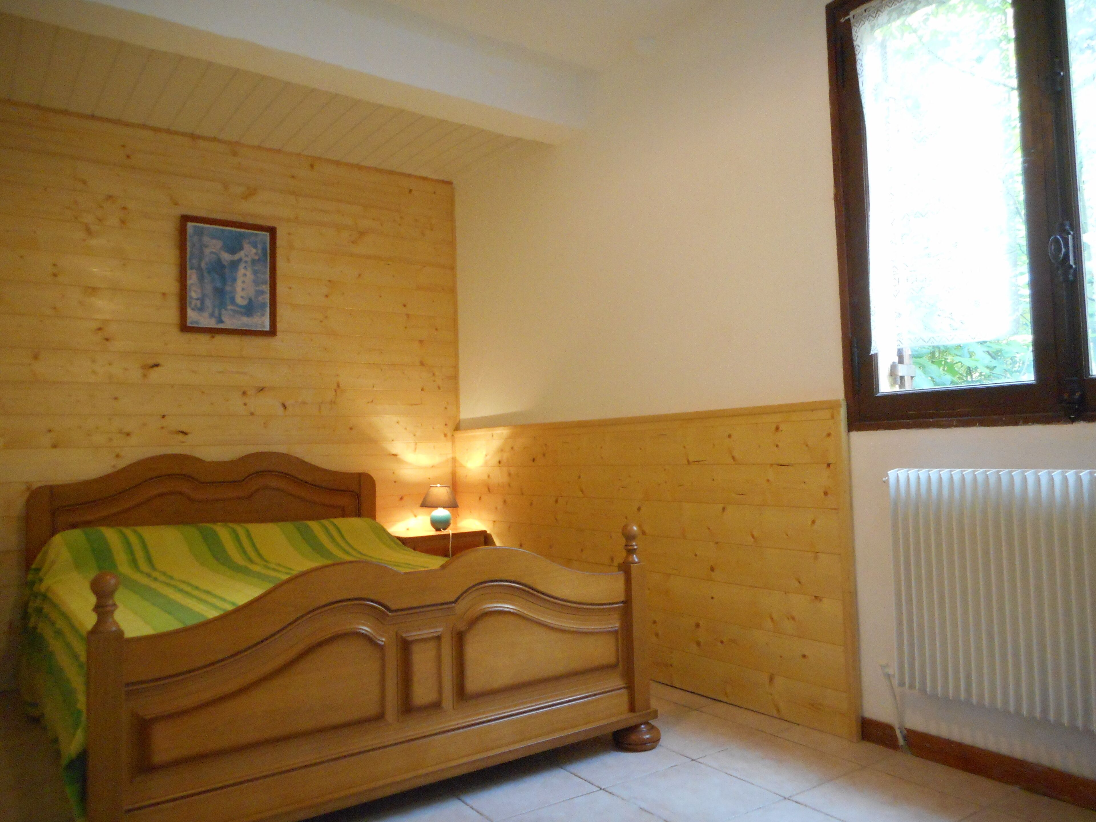 A wooden-framed bed with a green and yellow striped blanket, a bedside table with a lamp, and a window with blinds.