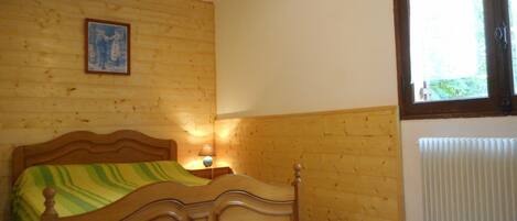 A wooden-framed bed with a green and yellow striped blanket, a bedside table with a lamp, and a window with blinds.