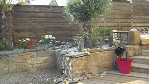 Un jardín con terraza de madera, pared de piedra y diversas plantas, incluyendo una palmera y arbustos florecientes.