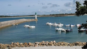 Escena costera con un faro, agua tranquila y varios barcos amarrados cerca de una costa rocosa.