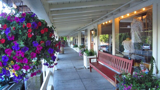 A covered outdoor area with hanging flower baskets, a red bench, and a storefront with mannequins.