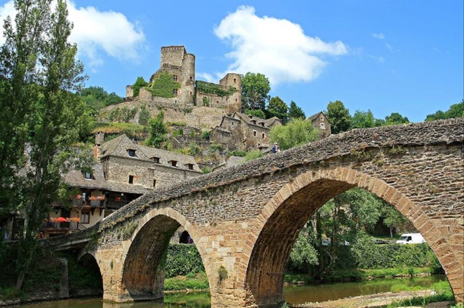 Eine Steinkonstruktion mit drei Bögen über einem Fluss, die zu einem historischen Steinhaus mit mehreren Ebenen und einem Turm auf einem Hügel führt.
