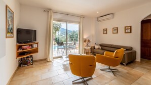 A living room with a TV, sofa, and two yellow chairs, a balcony with a table and chairs, and a view of greenery and buildings. - CASA VERDE - VILLA **** - Heated swimming pool - Near L'Ile-Rousse beach (Monticello)