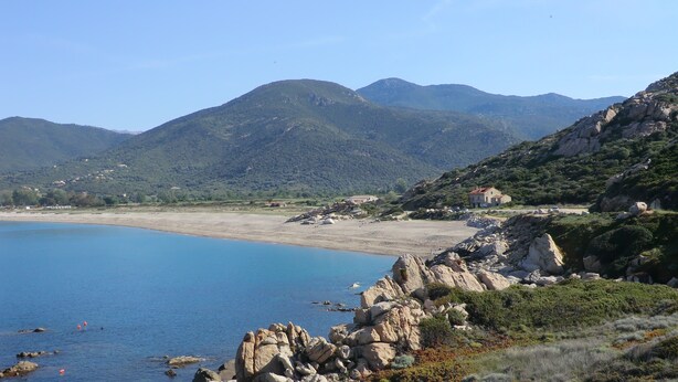 Paesaggio costiero con terreno roccioso, spiaggia e montagne in lontananza.