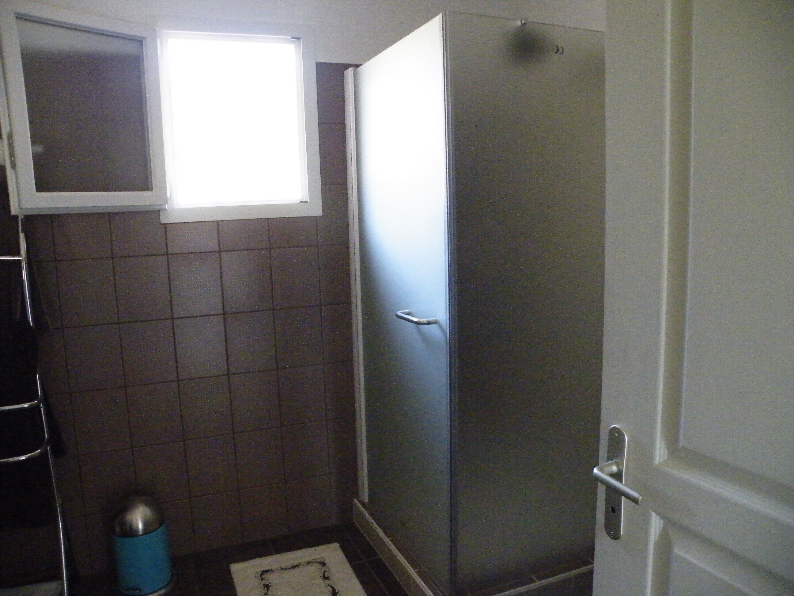 A small bathroom with a window, a tiled wall, a door, and a trash bin.
