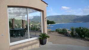 Un edificio con una porta vetrata che si apre su un patio con tavolo e sedie, affacciato su un paesaggio costiero con montagne e un corpo d'acqua.