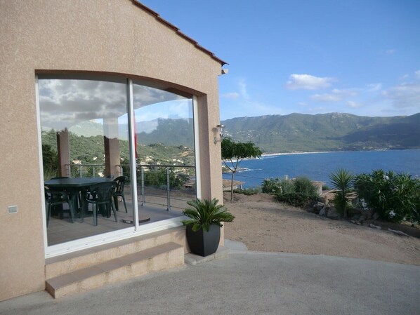 A building with a glass door leading to a patio with a table and chairs, overlooking a coastal landscape with mountains and a body of water. - VILLA - TIUCCIA- TERRA NOSTRA. (Casaglione)