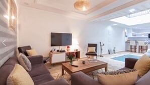 A modern living room with a sofa set, a coffee table, a TV, and a pool area in the background. - Riad Melilo heated swimming pool 2 steps from Jeema el Fna square (Marrakech)