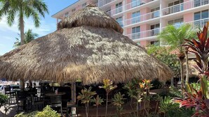 A thatched-roof outdoor dining area with tables and chairs, surrounded by tropical plants and a palm tree. - Palm Beach Shores Ocean Front Resort, Singer Island One Bedroom ~ Valet Parking (Palm Beach Shores)