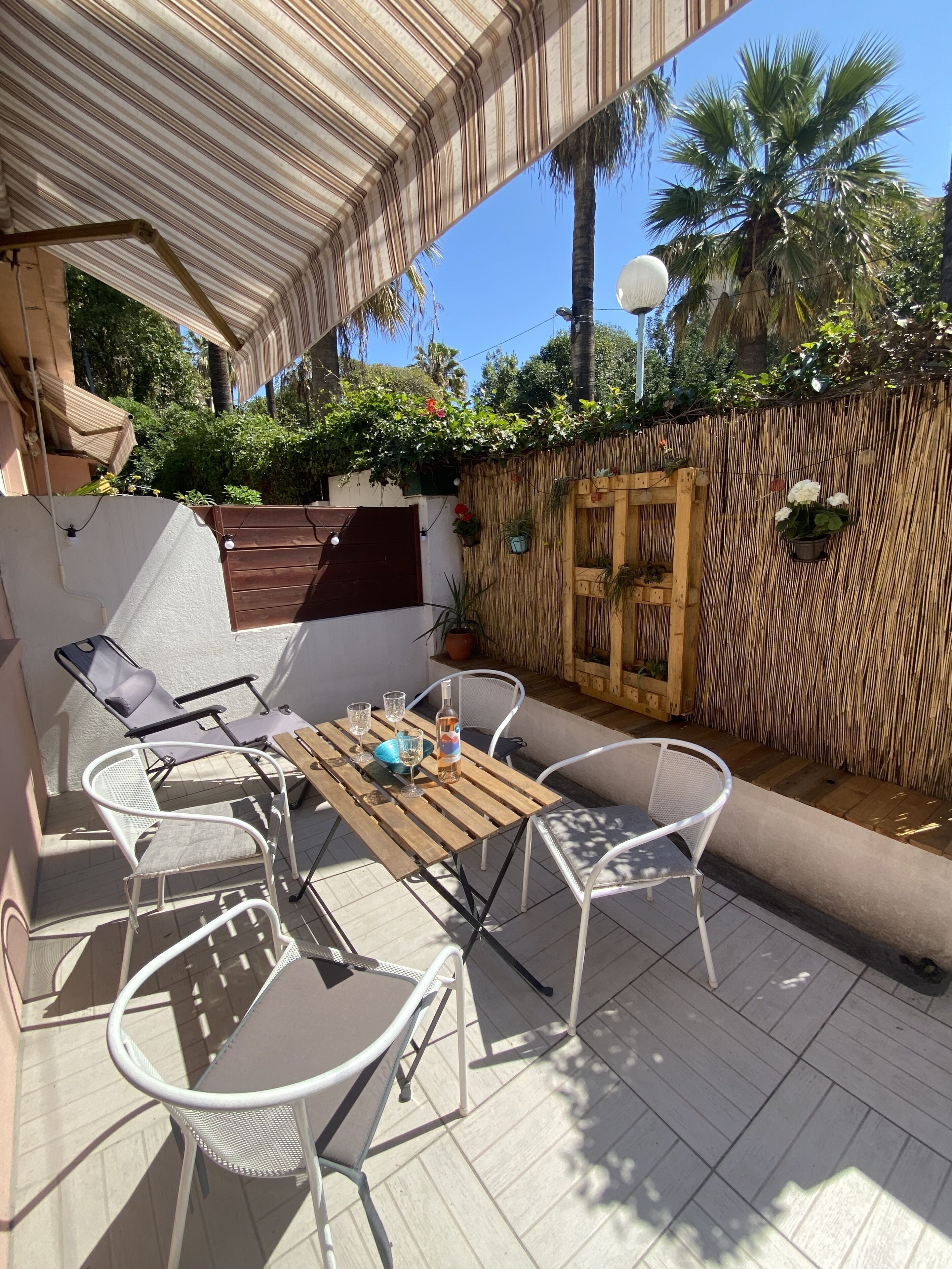 A patio with a wooden table and white metal chairs, surrounded by a bamboo fence and potted plants.