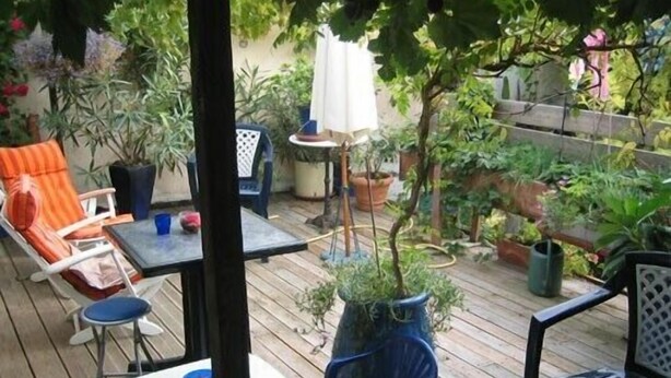 Una terraza de madera con mobiliario exterior, incluyendo sillas y una mesa, rodeada de macetas con plantas y un pequeño paraguas.