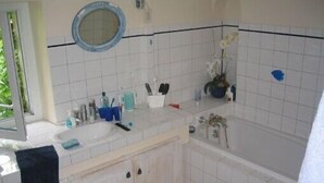 Un baño con lavamanos blanco, espejo y bañera. Hay artículos de tocador en la encimera y una toalla colgada en la pared.