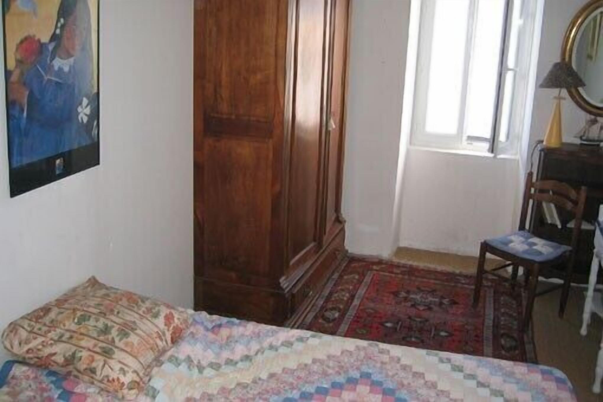 A bedroom with a bed, a wooden wardrobe, a chair, a desk with a lamp, and a window.